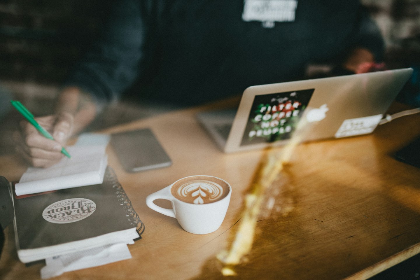 Person writing in notebook at a wooden table with laptop and coffee.
